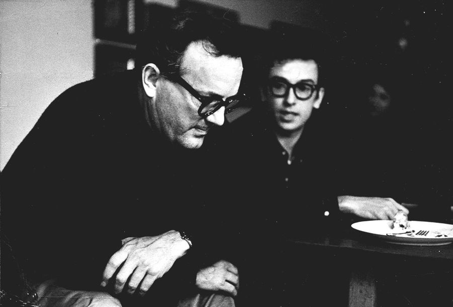 Two men at a table in a dim room. The older man in the foreground leans forward with his head lowered and his hands clasped over his knee; a younger man in heavy dark glasses sits behind him with a plate in front of him.
