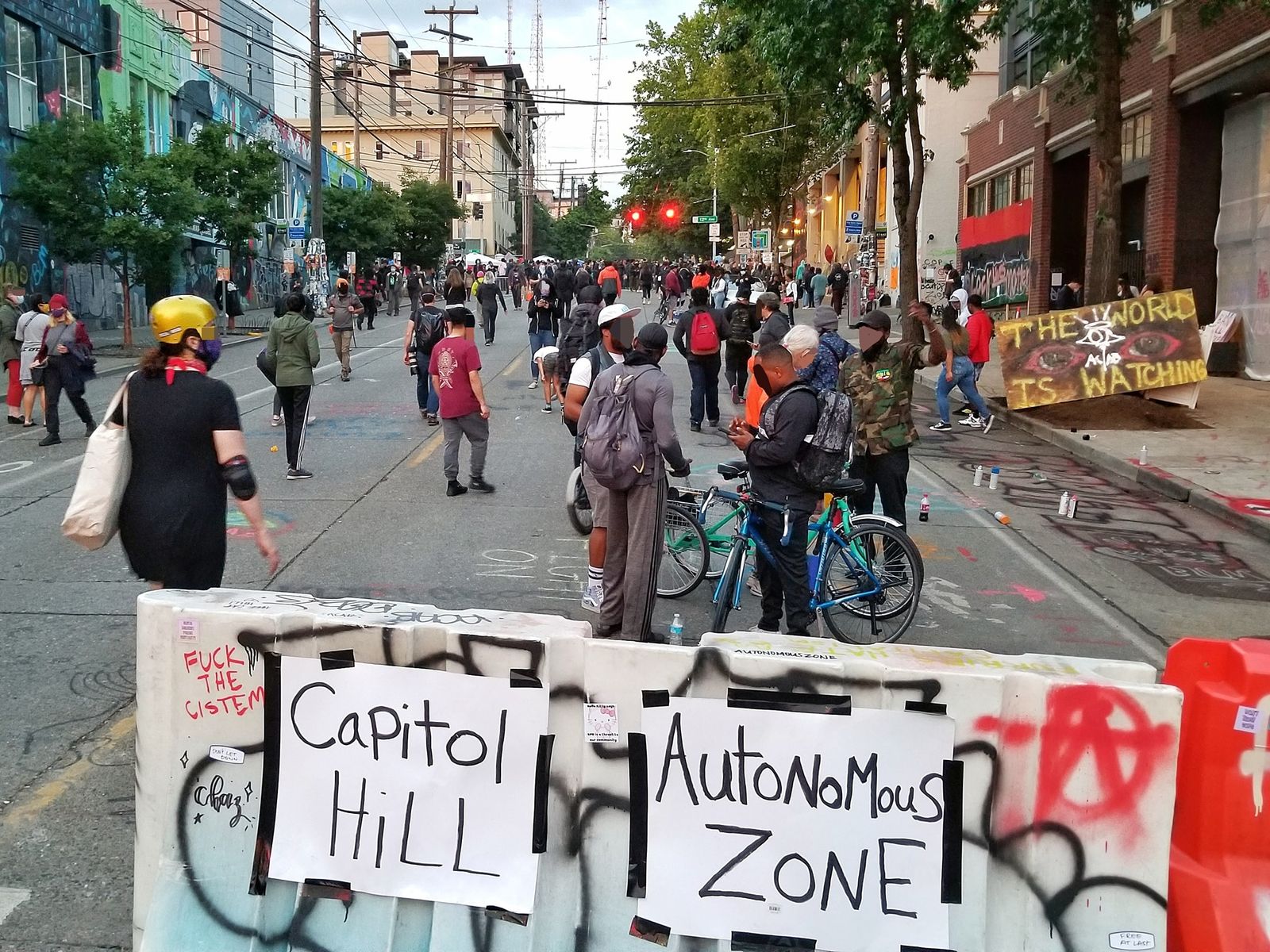 Two sheets of paper reading CAPITOL HILL and AUTONOMOUS ZONE are taped to a graffitied concrete highway barrier blocking a city street. Beyond it people stand and walk in the roadway between shopfronts, several of their faces blacked out by the photographer, and a hand-painted board propped on the pavement reads THE WORLD IS WATCHING.