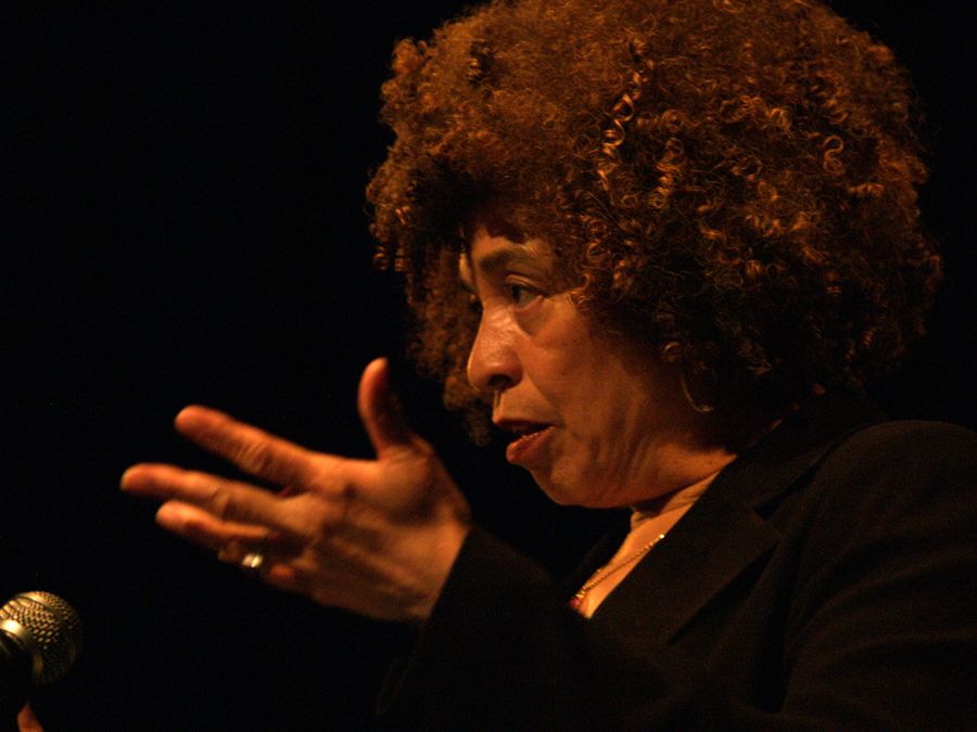 Angela Davis in profile against a black background, one hand raised mid-gesture as she speaks into a microphone.