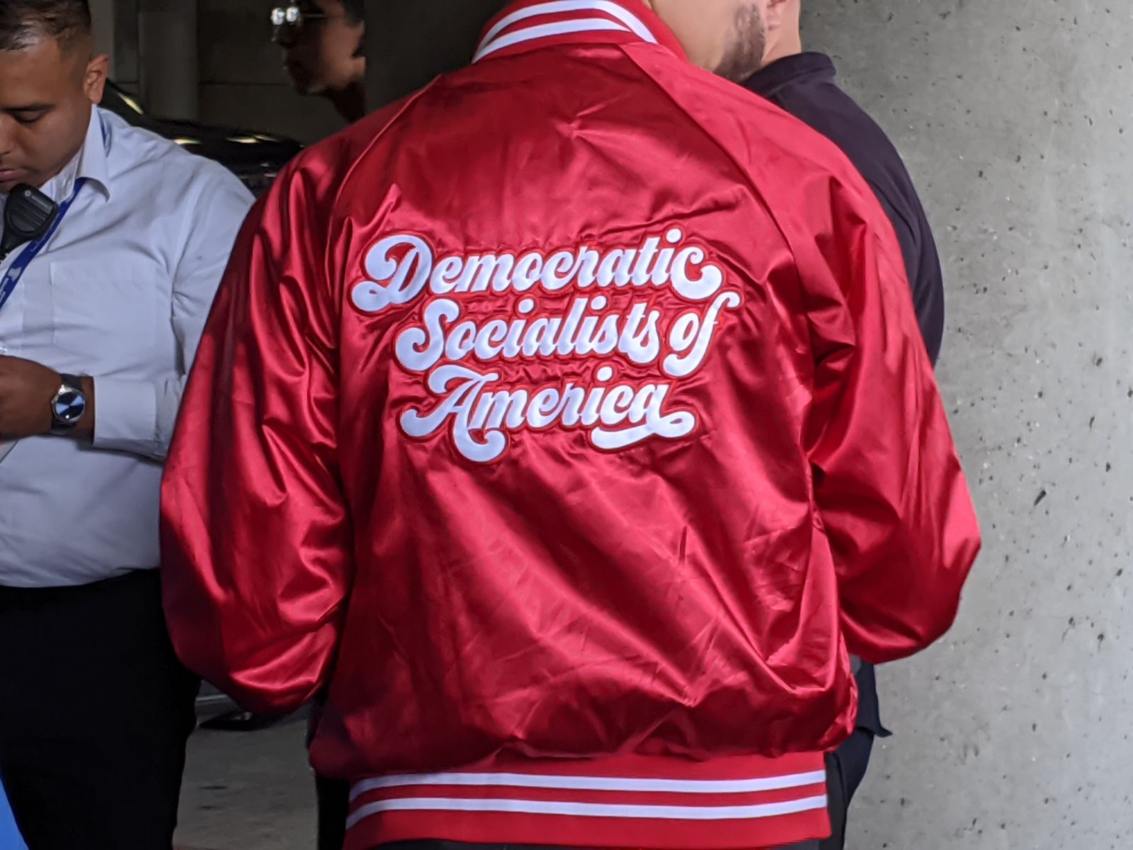 The back of a red satin jacket lettered in white script with the words Democratic Socialists of America, worn by a person standing with their back to the camera; other people stand out of focus on either side.