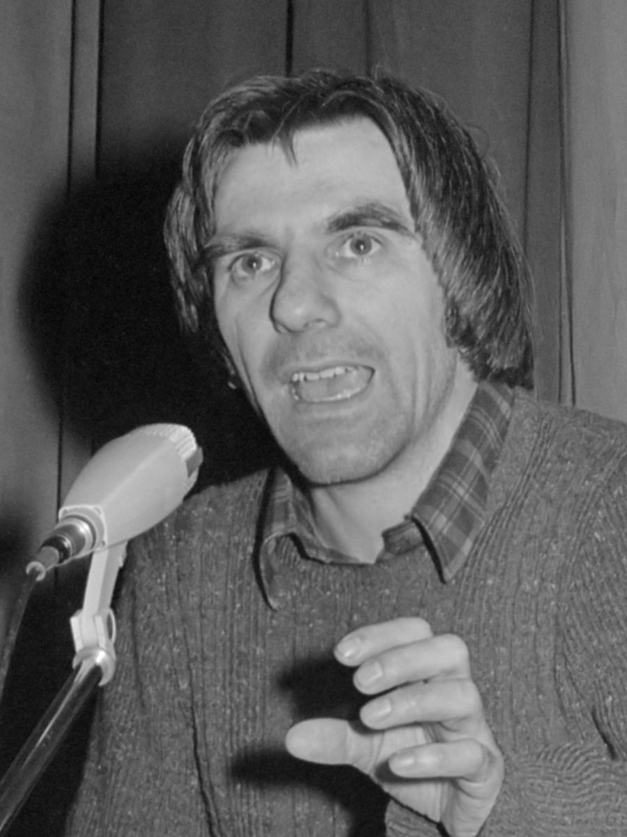 A man with long dark hair speaks into a table microphone, his mouth open mid-word and one hand raised in front of his chest.