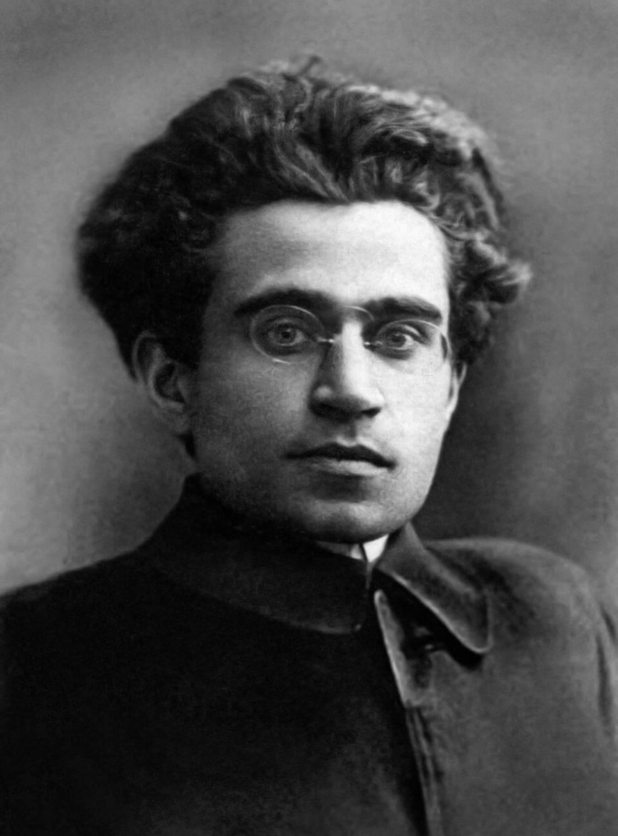 Head and shoulders portrait of a young man with a thick shock of dark hair, round wire-rimmed glasses and a heavy dark coat, facing the camera.