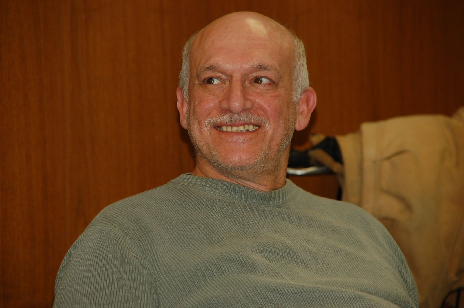 Mike Klonsky, a bald man with a grey moustache in a pale green sweater, turned to one side and smiling as he looks off camera against a wood-panelled wall.